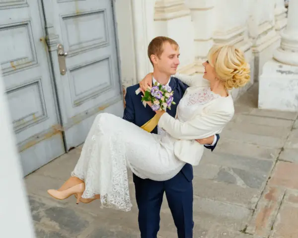 Pan młody niesie pannę młodą na rękach. Zastanawiasz się nad skutkami spóźnienia się z rezerwacją fotografa? Dlaczego 18 miesięcy to minimum?
