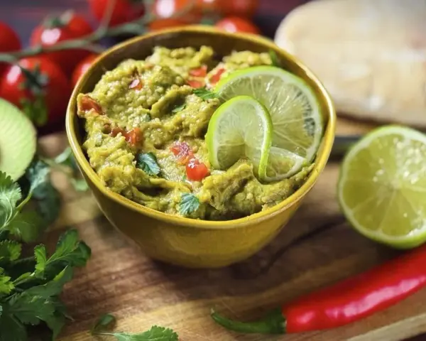 Frisches Guacamole-Rezept in einer gelben Schüssel, garniert mit Limettenscheiben. Daneben liegen Avocado, Koriander, Tomaten und Chili.