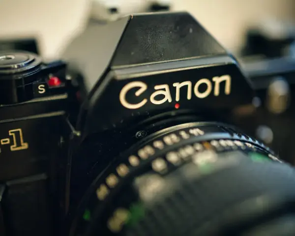 Zbliżenie na czarną lustrzankę Canon AE-1. Widoczny napis "Canon" i fragment obiektywu. Idealny do połączenia z aparatem.