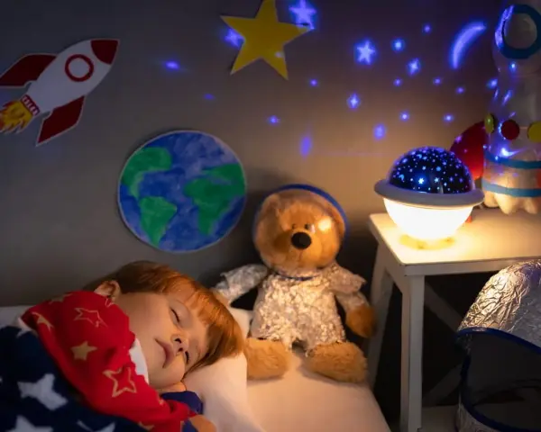 Śpiące dziecko pod kocem w gwiazdki, obok pluszowy miś w stroju astronauty. Na ścianie rakieta i Ziemia, a na suficie projektor dla dzieci na sufit wyświetla gwiazdy.