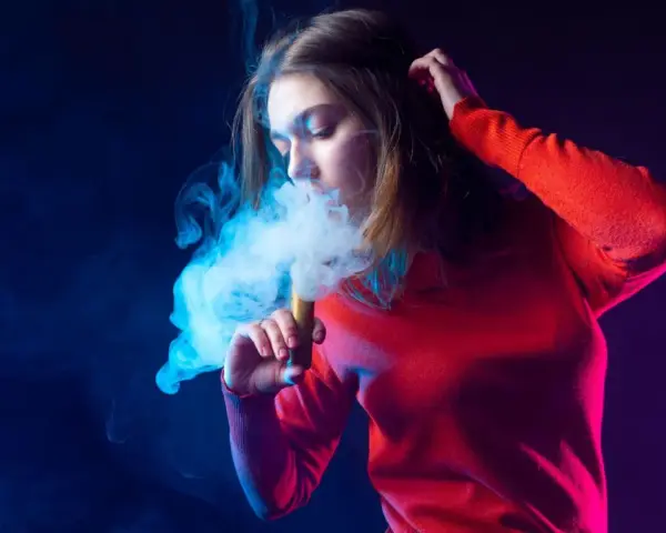 Joven exhala humo de un vaper, rodeada de luces azules y moradas.