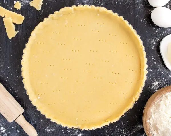 Ein vorbereiteter Mürbeteigboden in einer Tarteform, bereit für das 1 2 3 Teig Rezept. Zutaten wie Eier, Butter und Mehl sind daneben zu sehen.