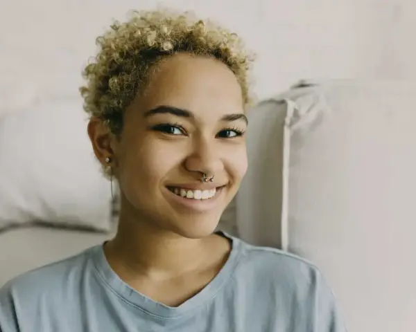 Uśmiechnięta młoda kobieta z kręconymi blond włosami i kolczykiem w nosie (septum piercing).