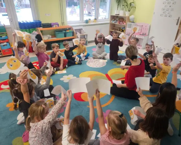 Wychowawca prowadzi zajęcia z dziećmi, które trzymają w rękach rysunki buziek wyrażających różne emocje.