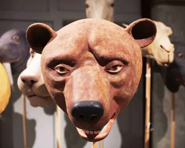 En brun björnmans ansikte, gjord av papier mache lim, med andra djurmasker i bakgrunden.