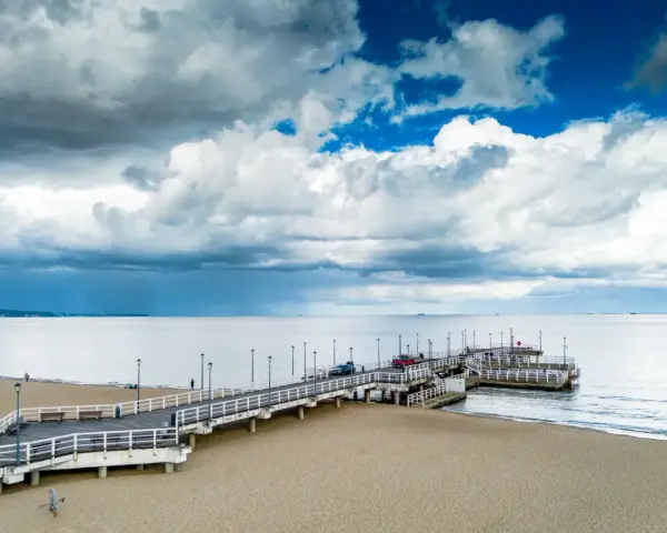 Długie molo w Brzeźnie wchodzi w spokojne morze. Na piaszczystej plaży widać kilka osób i samochody na molo.