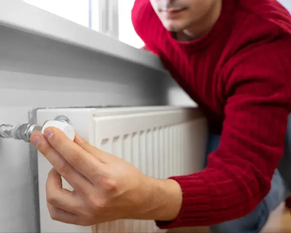 Mężczyzna w czerwonym swetrze reguluje termostat grzejnika, oszczędzając energię. To jego sposób na wykorzystanie bonu ciepłowniczy.