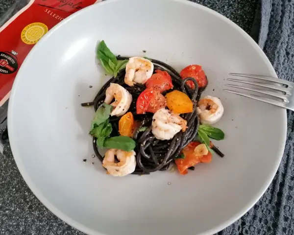 Czarny makaron z krewetkami, pomidorkami i bazylią na talerzu. Obok widoczna paczka spaghetti nero di seppia.