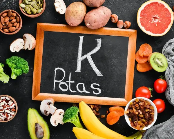 Tablica z napisem "K Potas" otoczona owocami i warzywami bogatymi w potas, jak banany, awokado, pomidory, ziemniaki, brokuły, rodzynki, morele, kiwi, migdały, pestki dyni. Pomaga zapobiegać hipokaliemii.