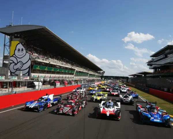 Una fila de coches de carreras listos para comenzar las 24 horas de Le Mans. El icónico muñeco de Michelin preside la tribuna.