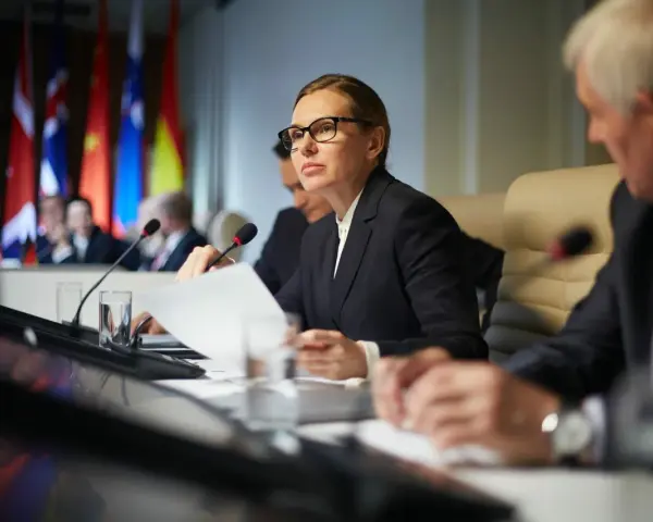 Kobieta w okularach analizuje dokumenty podczas konferencji międzynarodowej, gdzie omawiane są zagadnienia politologii.
