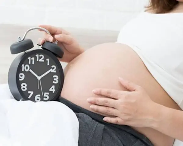 Zbliża się termin porodu. Kobieta trzyma dłoń na brzuchu obok budzika.