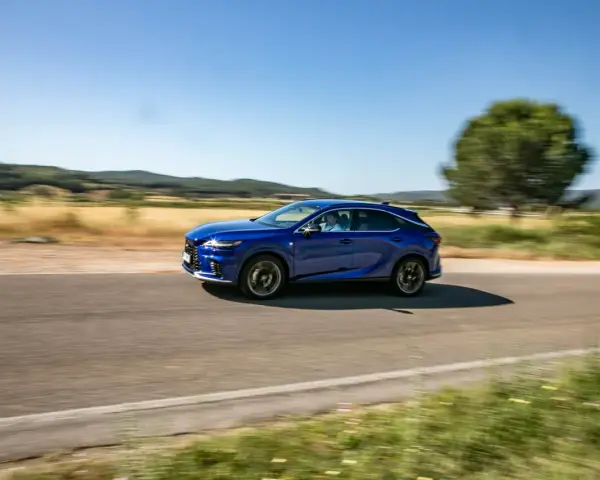 Niebieski Lexus 450h pędzi po wiejskiej drodze, otoczony zielenią i wzgórzami.