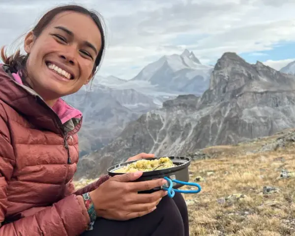 Sourire radieux d'une randonneuse savourant son repas en pleine montagne, avec des sommets enneigés en arrière-plan. Un moment de pur bonheur pour ce repas randonnée.