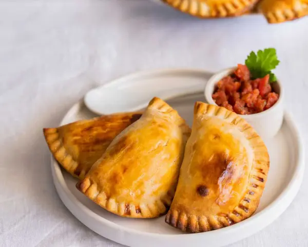 Tres empanadillas al horno doradas y crujientes, servidas en un plato blanco con un pequeño cuenco de salsa de tomate y una hoja de perejil.
