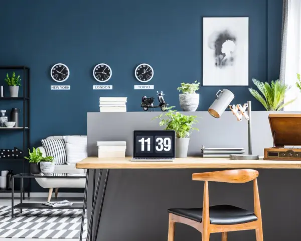 Biuro z zegarami pokazującymi czas w Nowym Jorku, Londynie i Tokio na ścianie w kolorze navy. Na biurku laptop, lampa i gramofon.