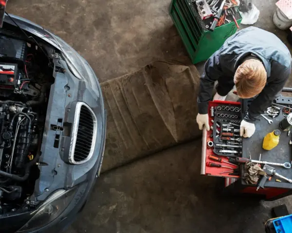 Meccanico con guanti bianchi esamina un motore sbiellato, circondato da attrezzi e un'auto con il cofano aperto.