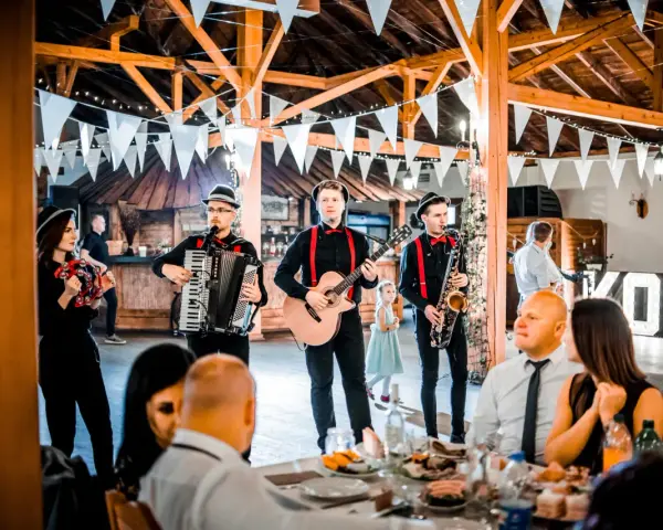 Zespół grający na weselu, z akordeonem, gitarą i saksofonem. W tle widać gości i dekoracje. Idealni do oprawy muzycznej imprez, znają teksty piosenek biesiadnych i weselnych.