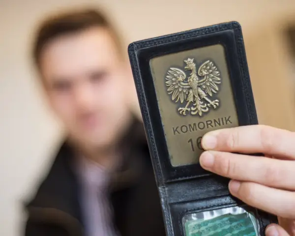 Komornik pokazuje legitymację z orłem. W tle niewyraźna postać mężczyzny, być może w związku z długiem osób prywatnych.