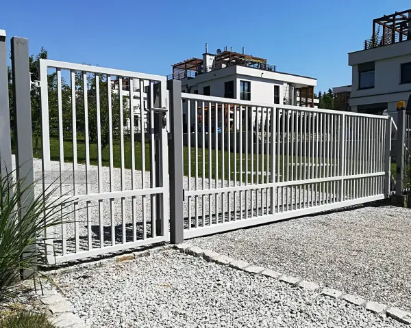 Brama z furtką bez słupka, otwarta na posesję z nowoczesnymi budynkami.