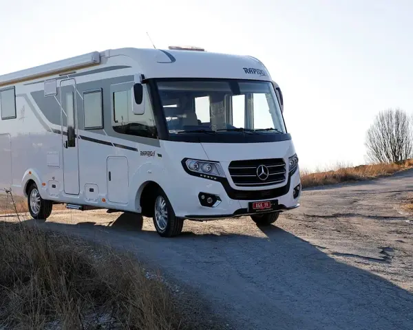 Vit husbil, en Rapido M66, parkerad på en grusväg med torrt gräs och träd i bakgrunden.