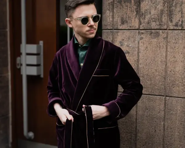 Elegancki mężczyzna w bordowej bonżurce z aksamitu, w okularach przeciwsłonecznych, stoi przy kamiennej ścianie.