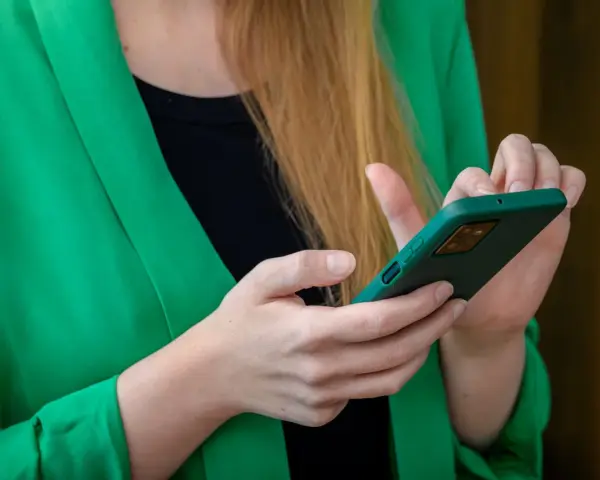 Kobieta w zielonej marynarce trzyma telefon. Szuka informacji, gdzie jest schowek w telefonie, aby ukryć ważne dane.