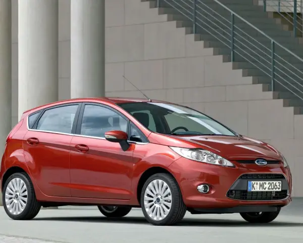 Czerwony Ford Fiesta MK6 zaparkowany przed nowoczesnym budynkiem z betonowymi kolumnami i schodami.