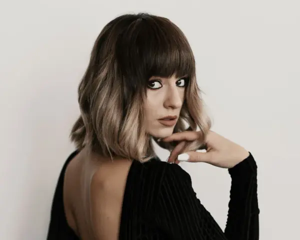Kobieta z modnym, **blunt bob** w odcieniach brązu i blondu, z grzywką, w czarnej sukience z odkrytymi plecami.
