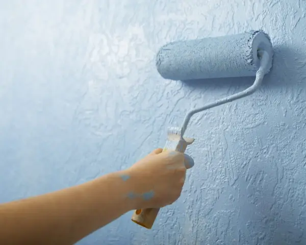 Mano aplica imprimación azul con rodillo a pared texturizada. La imprimación es el primer paso para un acabado perfecto.