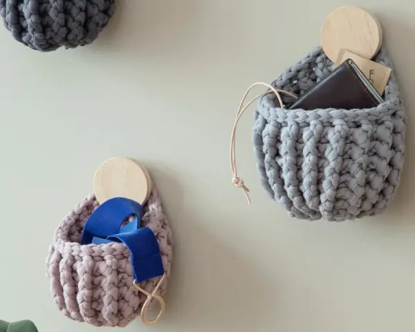 Three crocheted wall pockets in grey and pink, one holding a wallet, another blue ribbon, on a light wall.