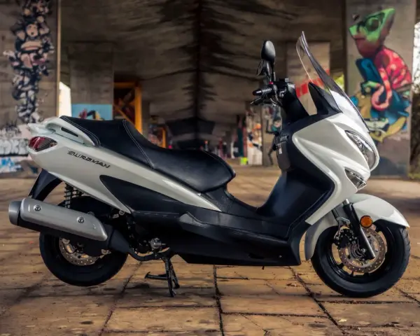 Biały skuter Suzuki Burgman 125, z czarnym siedzeniem, zaparkowany pod mostem z kolorowymi graffiti. Idealny do miasta, sprawdź suzuki burgman 125 dane techniczne.