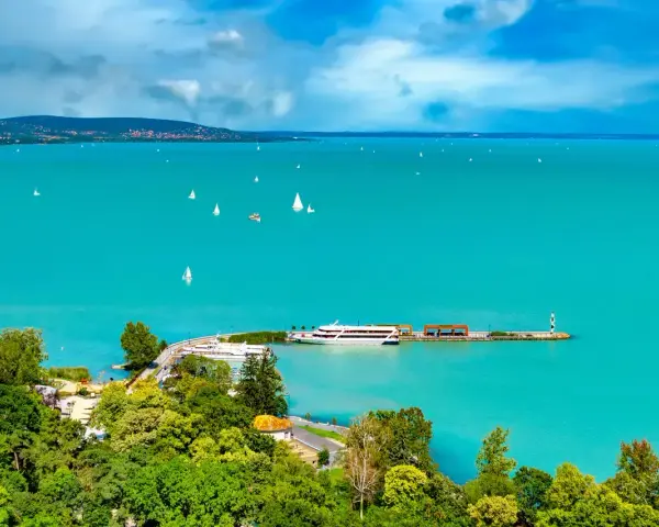 Turkusowe wody jeziora Balaton, pełne żaglówek i statków, z zielonymi wzgórzami w tle.