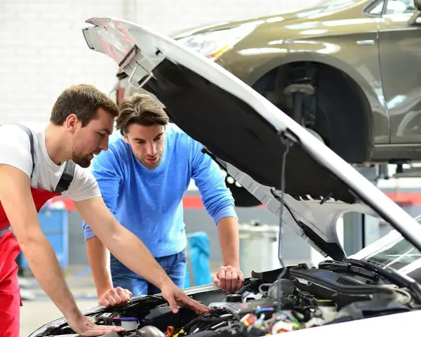 Zwei Männer blicken in den offenen Motorraum eines Autos. Der Mechaniker in Rot erklärt dem Kunden, welche Werkstatt mitgebrachte Teile einbaut.