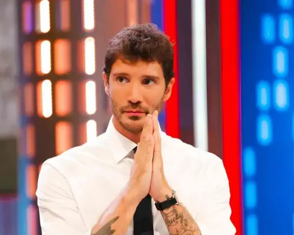 Santiago de Martino, con le mani giunte in preghiera, guarda intensamente verso l'obiettivo, con uno sfondo di luci colorate.
