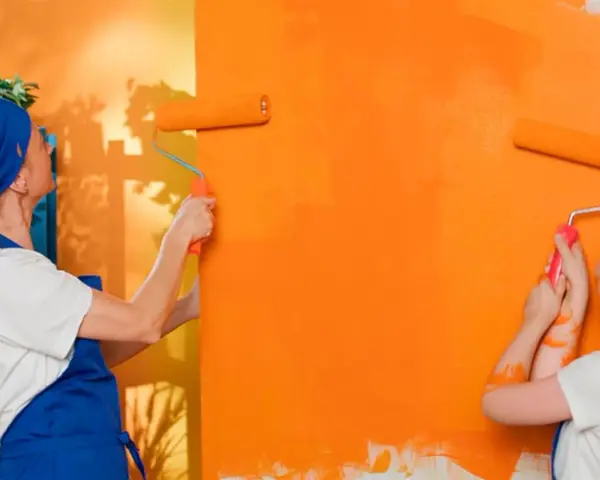 Dos personas pintan una pared de naranja brillante con rodillos. La pintura mural exterior cobra vida con vibrantes tonos anaranjados.