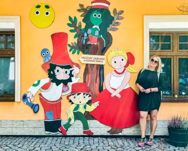 Kobieta pozuje przy kolorowej ścianie Muzeum Zabawek w Kudowie-Zdroju, Dolny Śląsk. Wesołe postacie z bajek towarzyszą jej w radosnej scenerii.
