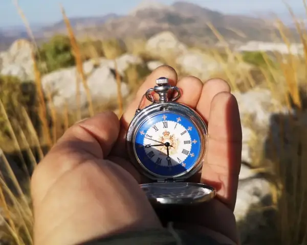 Mano sostiene un reloj de bolsillo antiguo con esfera azul y números romanos, útil para orientarse con reloj de agujas en un paisaje montañoso.