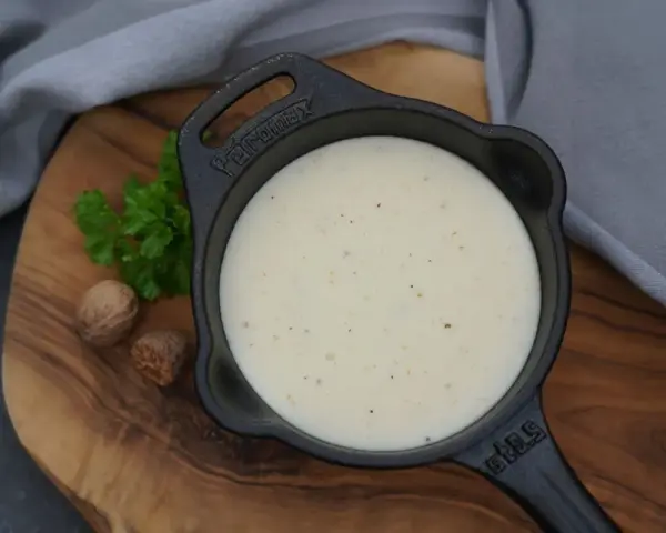 Cremige Bechamelsauce in einer Gusseisenpfanne, bereit für die Lasagne. Muskatnuss und Petersilie deuten auf die feinen Aromen hin.