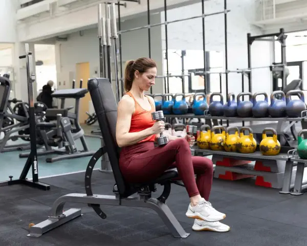 Kvinna utför axelpress med hantlar på en träningsbänk i ett gym.
