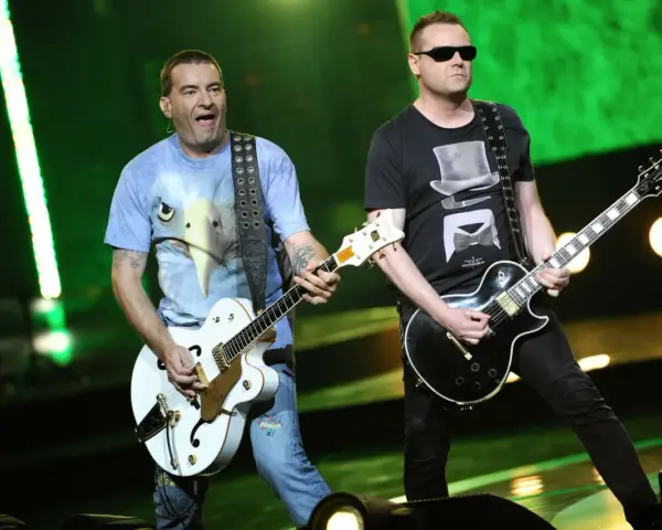 Dwóch członków Kobranocki gra na gitarach podczas koncertu. Jeden w koszulce z orłem, drugi w czarnej z cylindrem i muszką.