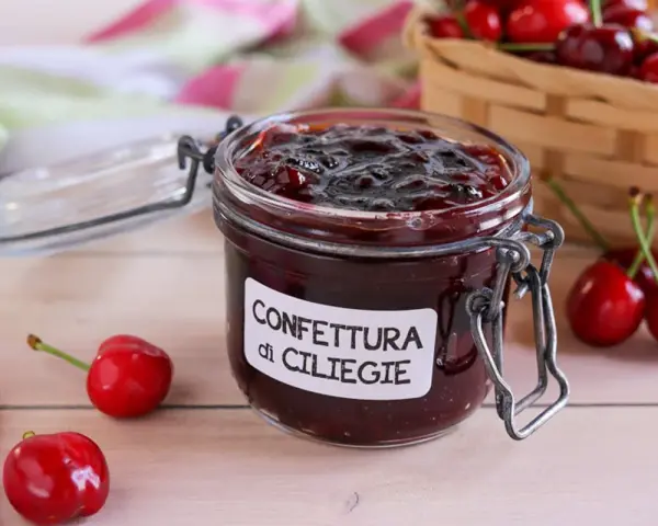 Un vasetto di marmellata di ciliegie fatta in casa, con ciliegie fresche e un cestino sullo sfondo.