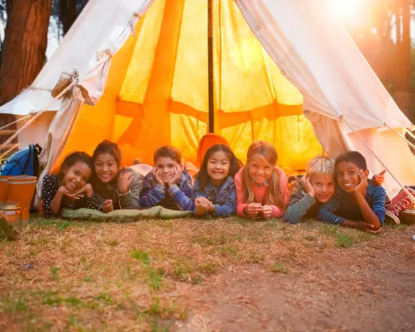 Sept enfants souriants allongés devant une tente de camping éclairée par le soleil.