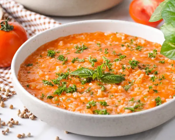 Eine cremige Tomatensuppe mit Reis, garniert mit frischer Petersilie und Basilikum. Daneben liegen frische Tomaten und Gewürzkörner.