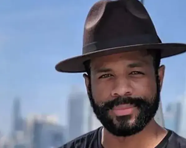 Hombre con sombrero y barba espesa, sonriendo ante un fondo de edificios y cielo azul.