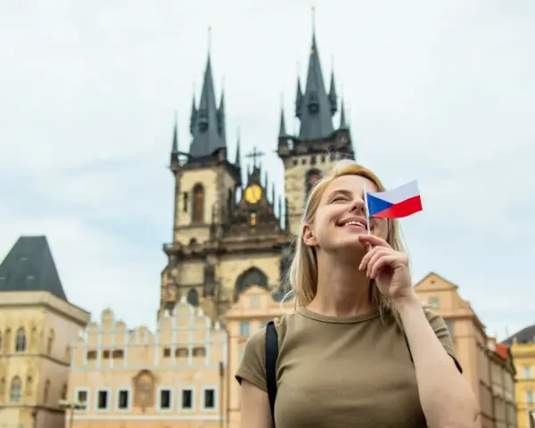 Uśmiechnięta blondynka z flagą Czech zasłaniającą oko, marzy o wysokich zarobkach w Czechach. W tle praskie kamienice i kościół.