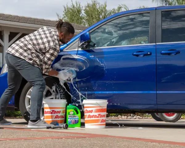 A man washes a blue car in the sun, with buckets and car wash soap nearby. It's a good day to wax a car in the sun.