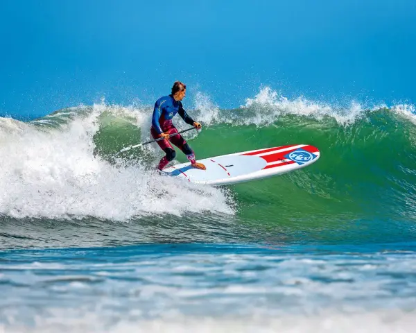 Ein Mann im Neoprenanzug surft auf einem SUP-Board eine grüne Welle. Das SUP-Wellenreiten erfordert Balance und Geschick.