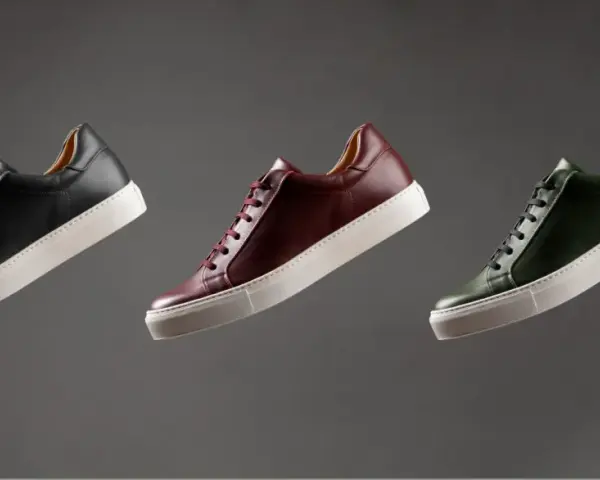 Sneakers marche famose in pelle nera, bordeaux e verde, con suola bianca, fluttuano su sfondo grigio.