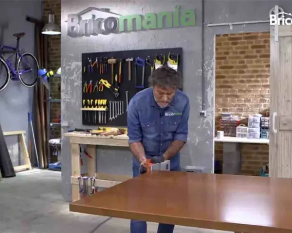 Hombre lijando mesa de madera. El taller tiene herramientas colgadas y una bicicleta morada. La madera hinchada por humedad se repara.